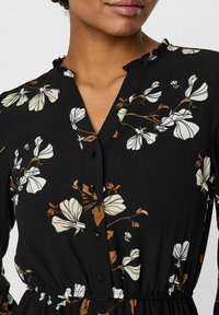 Musta mekko kukkakuviolla, jossa on valkoisia ja ruskeita kukkia, napitus ja röyhelökaulus. Teksturoitu kangas, jossa on kapea vyötärö.