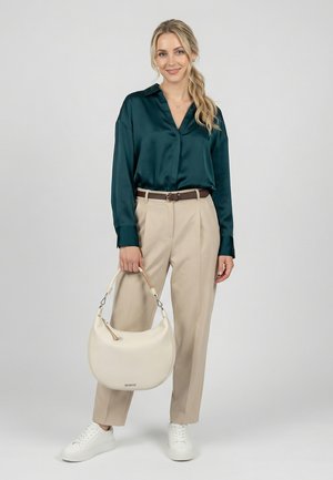 Frau in dunkelgrüner Satinbluse, beiger Hose, weißen Sneakers, die eine weiße Handtasche mit gebogenem Griff vor grauem Hintergrund hält.