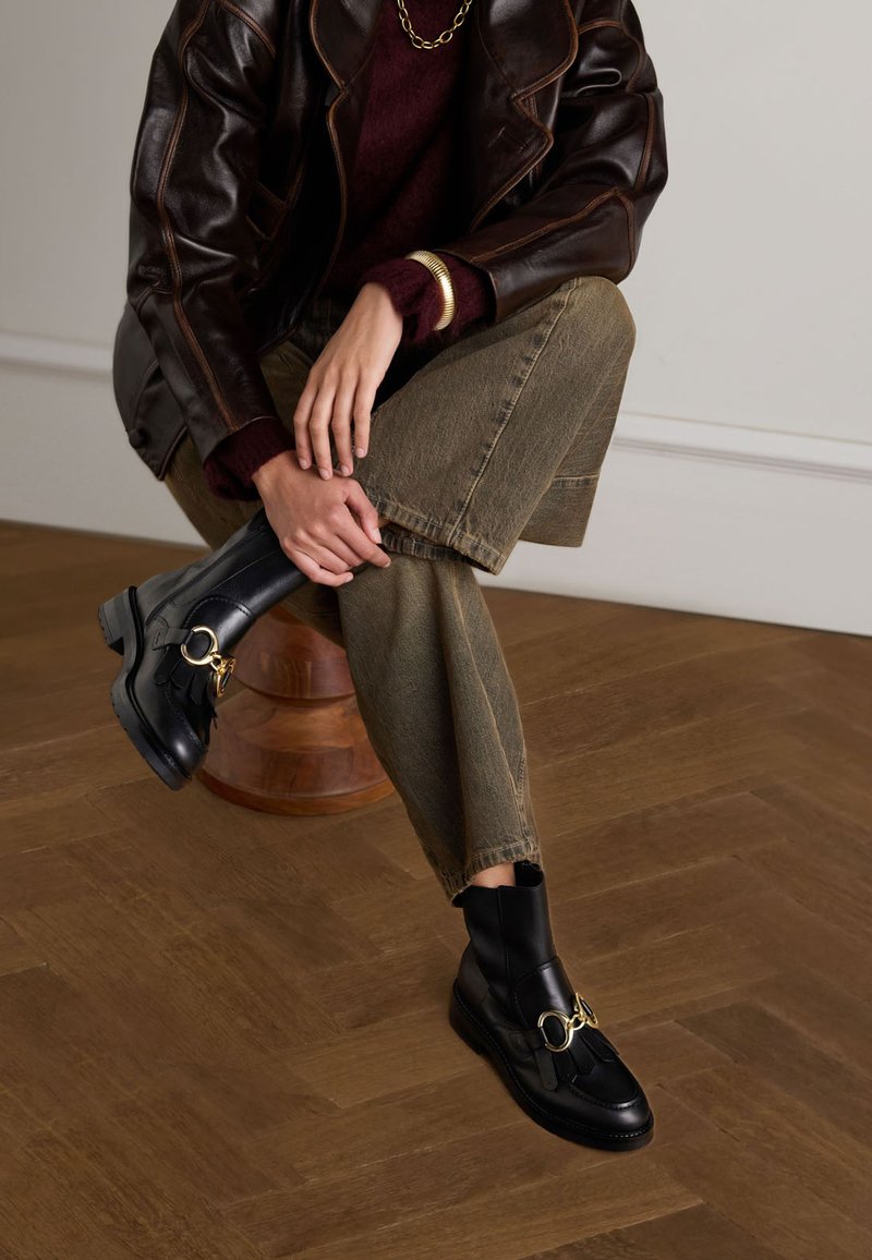 Bottines en cuir noir avec des accessoires en or et un gland décoratif. Portées avec un jean cropped vert olive et une veste en cuir marron foncé.