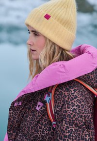 Prešívaná bunda s ružovou vonkajšou vrstvou a vnútornou podšívkou s hnedým leopardím vzorom, doplnená svetložltou pletenou čiapkou a batohom.