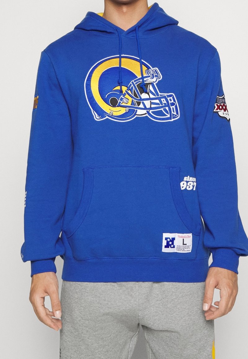 Sweat à capuche bleu avec un grand logo de casque de football jaune et blanc, écusson du Super Bowl sur la manche, poche avant, porté par une personne en pantalon gris.