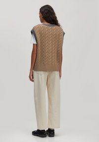 Beige kabelgebreide vest met korte mouwen, voorzien van een geribbelde ronde hals en zoom. Draagt over een wit T-shirt en gecombineerd met crèmekleurige wijde broek.