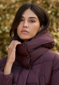 Gevoerde, gestikte jas in donker bordeaux met een hoge kraag, knoopsluitingen en een gestructureerde afwerking, voorzien van een diagonale quiltpatroon.