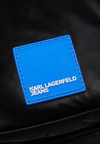 Črn tekstilni nahrbtnik z oznako iz modre gume v obliki kvadrata, na kateri je bele embosirane besede "KARL LAGERFELD JEANS", obročene z modrim šivom.