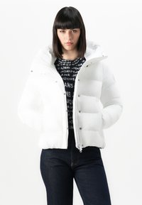 Witte pufferjas met een capuchon, voorzien van drukknoopjes en een rits. Eronder een gebreide zwarte trui, gecombineerd met donkere jeans.