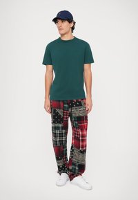 Zelené bavlněné tričko, volné kostkované kalhoty s černými, červenými a zelenými vzory, bílé tenisky a námořnická baseballová čepice.