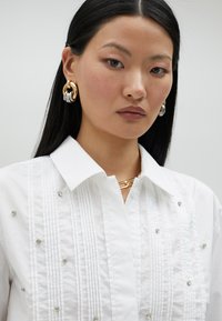 Chemise blanche à col classique avec des plis et des embellissements en cristal dispersés, associée à de grandes boucles d'oreilles en or et en argent et un collier en chaîne dorée.