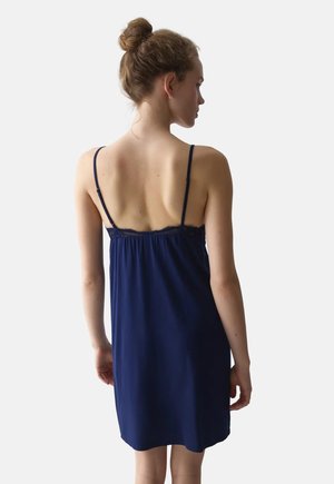 Femme avec les cheveux en chignon portant une robe sans manches bleu marine à fines bretelles, vue de dos sur un fond blanc uni.
