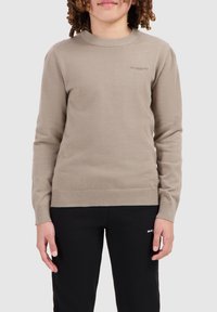 Beige gebreide trui met een ronde halslijn en geribbelde manchetten en zoom. Bevat subtiele branding op de borst. Gepaard met zwarte broek.