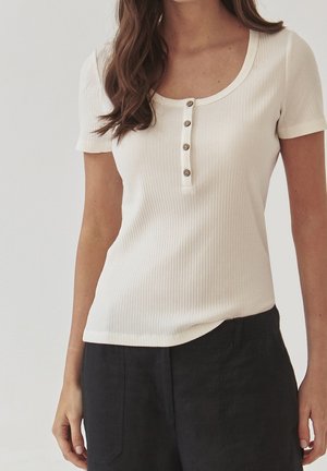 Femme portant un t-shirt crème côtelé à manches courtes avec quatre boutons et un pantalon noir ample sur un fond uni.