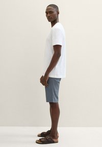 Weißes T-Shirt mit kurzen Ärmeln, kombiniert mit blauen Shorts, in einem lockeren Schnitt. Das Modell trägt dunkelbraune Sandalen mit zwei Schnallen.