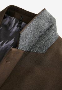 Ruskea kangastakki, jossa on harmaa huopakaulus. Sisäpuolella on kuviollinen vuori, jossa on tummia ja vaaleita sävyjä. Ulkopinnassa näkyviä ompelujälkiä.