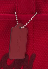 Sac en tissu rouge texturé avec une étiquette en cuir assortie embossée "COACH NEW YORK" suspendue à une chaîne à billes argentée.