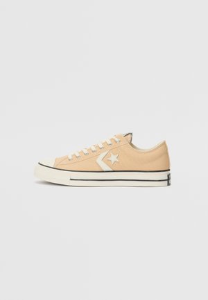 Basket basse beige avec étoile blanche et logo chevron, lacets blancs et semelle blanche avec rayures noires sur un fond uni.