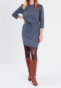 Kleid mit floralem Muster, tailliertem Schnitt, Dreiviertelärmel und knielangem Saum, kombiniert mit braunen knielangen Stiefeln.