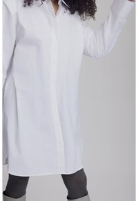 Camisa blanca de algodón con botones, con cuello puntiagudo y mangas largas. Dobladillo recto con un ajuste relajado. Se lleva sobre leggings grises.