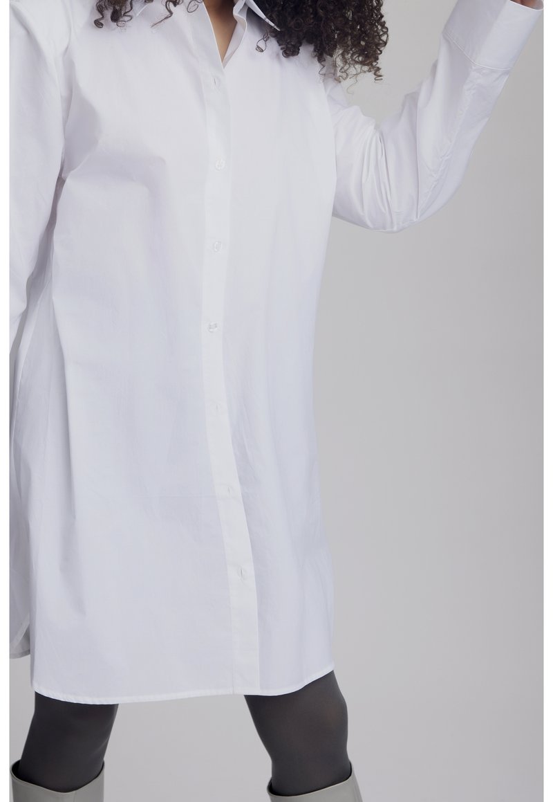 Camisa blanca de algodón con botones, con cuello puntiagudo y mangas largas. Dobladillo recto con un ajuste relajado. Se lleva sobre leggings grises.