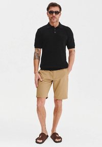 Czarna polo z krótkim rękawem, khaki szorty, brązowe sandały, z widocznymi tatuażami na rękach. Gładkie tło, osoba stoi w pozycji wyprostowanej.