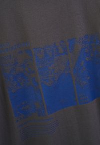 Koszulka z grafiką wykonana z ciemnoszarej tkaniny z niebieskim tekstem i wzorami przedstawiającymi mapy miast oraz hasła. Tekst zawiera "OFF DUTY GEAR."