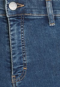 Mörkblå denimpanel med beige sömmar, en metallknapp med "TOPSHOP" och en sydd fickdetalj på sidan.