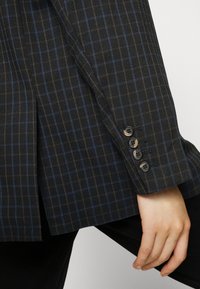 Černobílé kostkované sako s přizpůsobeným střihem, texturovanou tkaninou a čtyřmi černými knoflíky na manžetách.