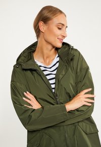 Jachetă verde impermeabilă cu glugă, închidere cu fermoar și buzunare mari. Pur tată peste o cămașă cu dungi, având o textură netedă și lucioasă.