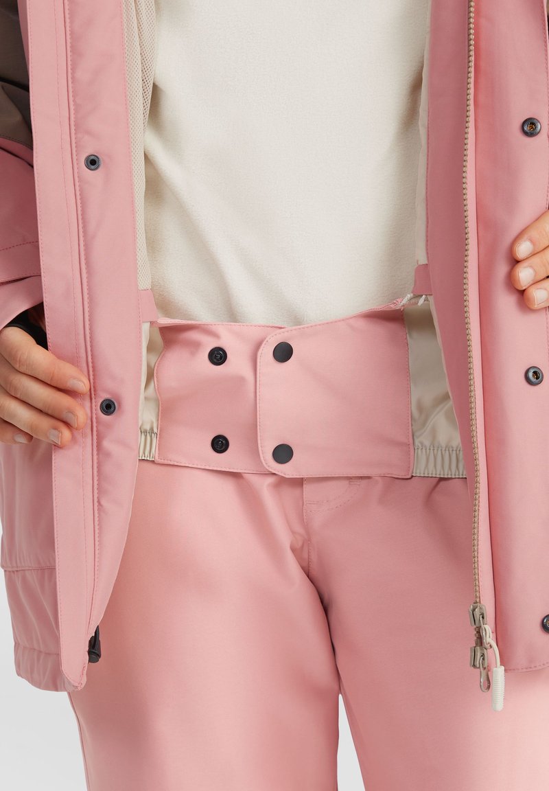 Roze outerwear bestaande uit een gevoerde jas en bijpassende hooggehakte broek. De tailleband bevat zwarte drukknoopjes en elastische details.