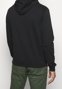 Felpa nera con cappuccio regolabile, polsini e vita a coste, abbinata a pantaloni verdi con una texture liscia e design minimale.
