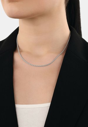 Collana in argento con design intrecciato, indossata su una maglietta chiara e sotto un blazer nero, che mette in mostra una superficie liscia e un profilo sottile.