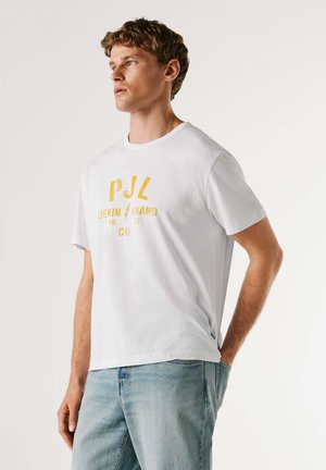 Valge puuvillane T-särk, millel on kollane graafiline trükk tekstiga "PJL DENIM BRAND 1913 CO.". Kaelus on ümara lõikega ja varrukad lühikesed. Vabaaja lõige.