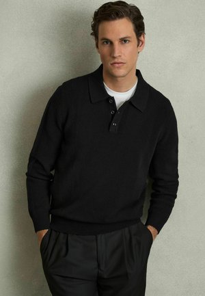 Pullover nero lavorato a maglia con colletto e chiusura a tre bottoni. Presenta una texture a coste sulle spalle e sui polsini. Indossato sopra una camicia bianca.