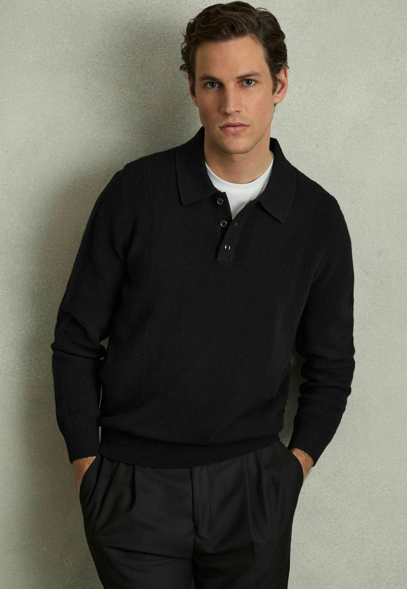 Pullover nero lavorato a maglia con colletto e chiusura a tre bottoni. Presenta una texture a coste sulle spalle e sui polsini. Indossato sopra una camicia bianca.