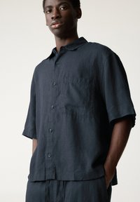 Camicia in lino scuro navy a maniche corte con colletto, taschino frontale e chiusura con bottoni, caratterizzata da una vestibilità rilassata e da una texture morbida.