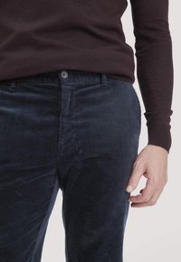 Marineblaue Cordhosen mit vertikaler Rippung, ausgestattet mit einem Knopfverschluss und vorderen Taschen. Getragen mit einem dunklen Pullover.