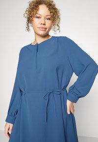 Vestido azul com mangas longas, punhos franzidos, decote redondo e um detalhe de atar na cintura. Feito de um tecido liso com detalhes de design mínimos.