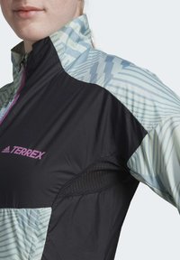 Schwarze und hellblaue Outdoor-Jacke mit hohem Kragen, versehen mit geometrischen Mustern und Mesh-Details, mit dem Markennamen "TERREX" in Pink.