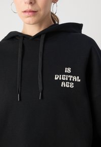 Svart hettegenser med en frontlomme, snorer og teksten "IS DIGITAL AGE" i en lysfarget, fet skrift.