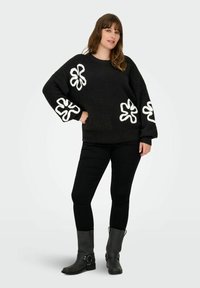 Zwarte gebreide trui met een losvallende pasvorm en witte bloemenappliqué op de mouwen. Gecombineerd met zwarte slim-fit broeken en grijze enkellaarzen.