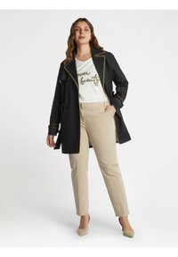 Svart trenchcoat med guldkant, vit skjorta med glittrig text och beige byxor. Korta klackar fulländar outfiten.