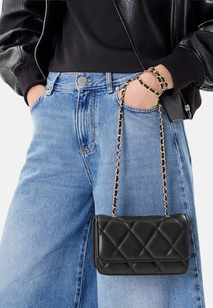 Frau trägt schwarze Lederjacke und schwarzes Oberteil mit hellblauen Jeans, hält eine gesteppte schwarze Handtasche mit goldener Kettenriemen am Handgelenk.