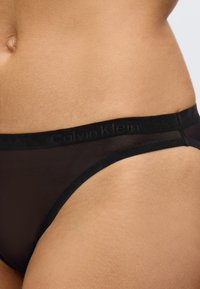 Juodas permatomas bikini stilius su lygiu juosmeniu, ant kurio iškilmingai išrašyta "Calvin Klein" logotipo. Minkšta medžiaga, minimalistiškas dizainas.