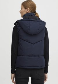 Marinblå puffärmlig väst med hög krage och huva, med horisontellt quiltade sektioner och sidodragkedjor, kombinerad med en svart långärmad tröja.