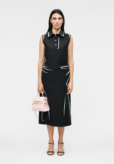 Γυναίκα που φοράει μαύρο αμάνικο μεσαίο φόρεμα με λευκές περιμετρικές λεπτομέρειες, κρατώντας μια μικρή ροζ τσάντα και μαύρα σανδάλια με λουράκι στον αστράγαλο.