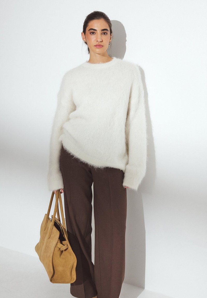 Un pull blanc duveteux à manches longues, associé à un pantalon tailleur marron. Le modèle porte un grand sac à main en daim tan.