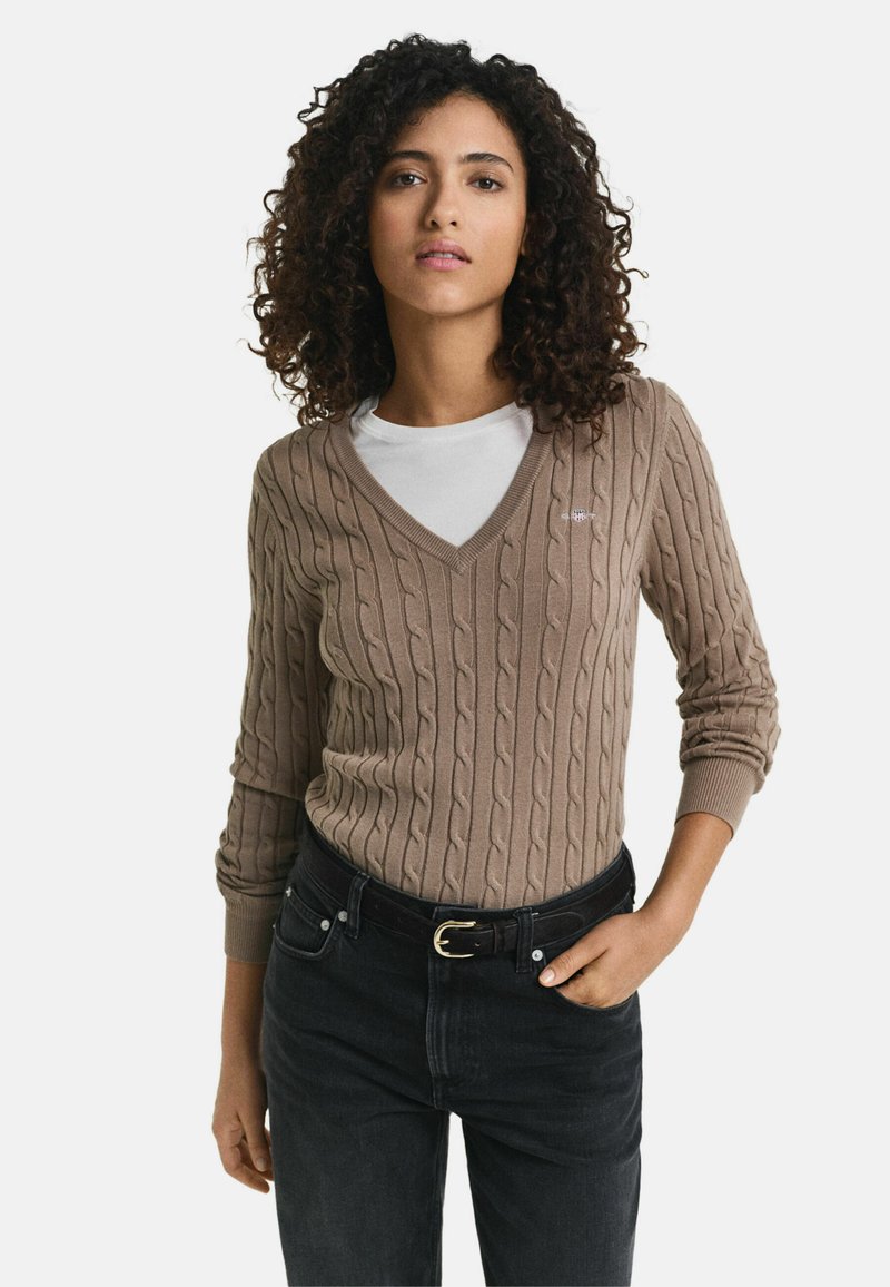 Beige Strickpullover mit V-Ausschnitt und langen Ärmeln, getragen über einem weißen Hemd; kombiniert mit hoch taillierten schwarzen Jeans und einem Gürtel.