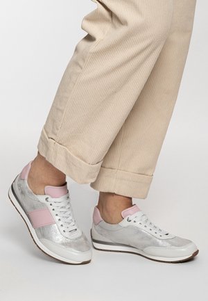 Füße in weißen und silbernen Sneakers mit rosa Akzenten, kombiniert mit beigen Cordhosen, an den Bündchen hochgekrempelt, vor schlichtem Hintergrund.