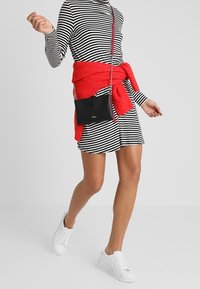 Černobílé pruhované šaty s dlouhými rukávy, spárované s červeným svetrm převázaným v pase a bílými teniskami. Součástí je černá crossbody taška.