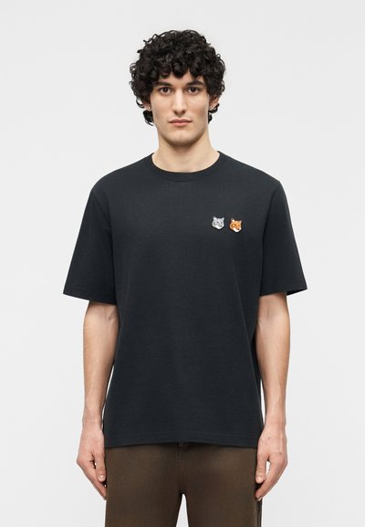 Jeune homme aux cheveux bouclés et foncés portant un T-shirt noir orné de petites écussons tête de loup et renard sur le côté gauche de la poitrine.