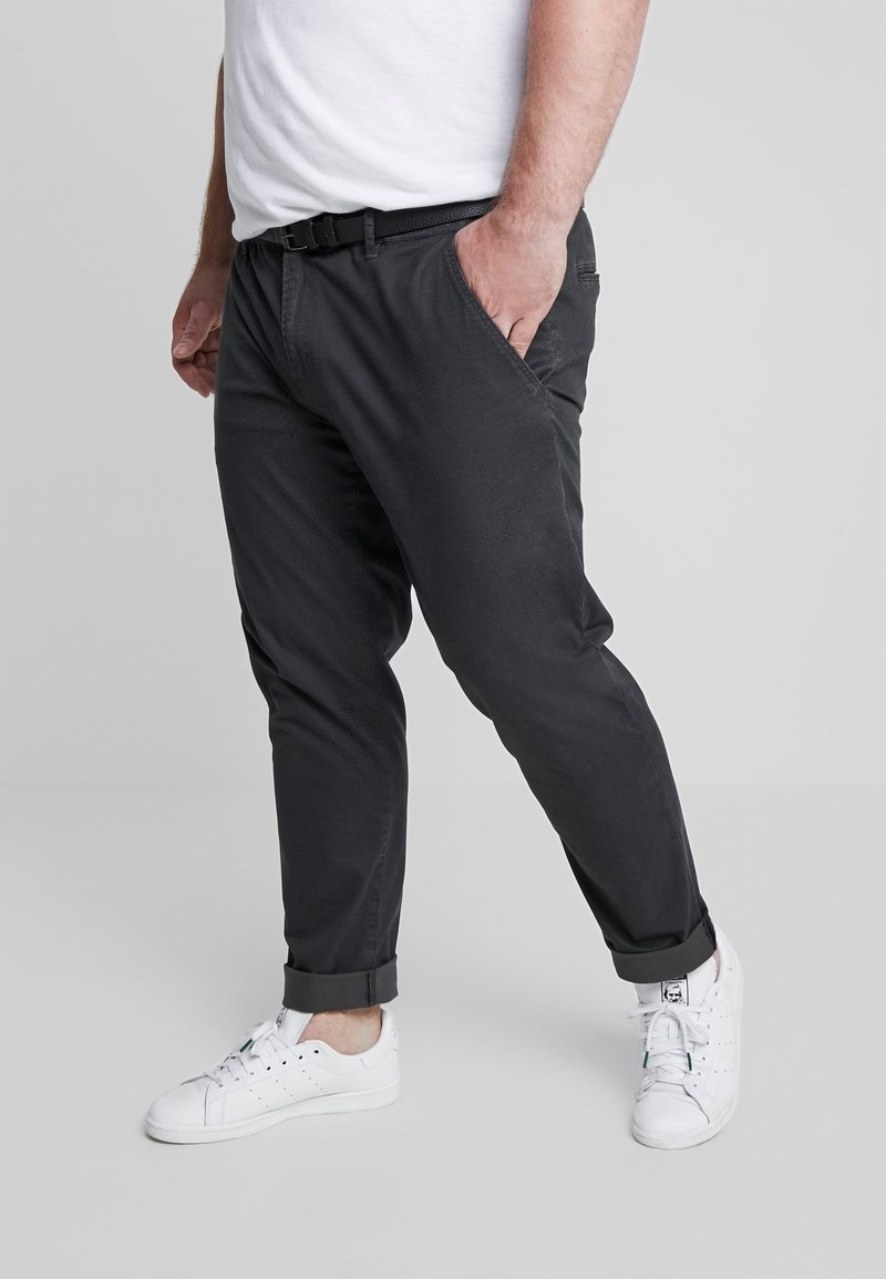 Homme portant un pantalon noir retroussé, une ceinture blanche, des baskets blanches et un t-shirt blanc, debout avec une main dans la poche, fond gris clair.