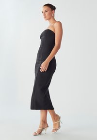 Žena v elegantních černých midi šatech bez ramínek a bílých sandálech na podpatcích stojí na světlém jednobarevném pozadí.